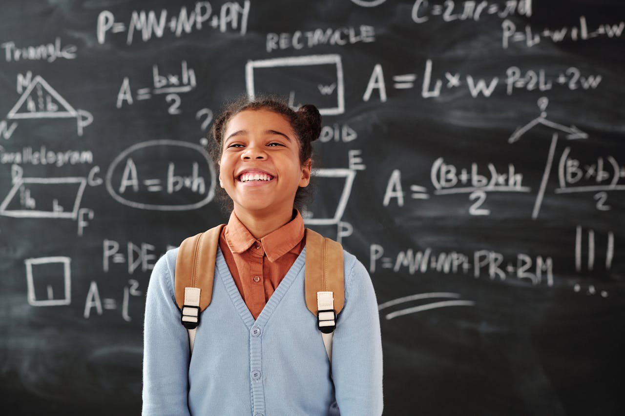 Smiling girl with backpack in a classroom, standing in front of a chalkboard with math formulas.