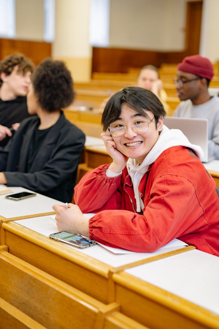 Asian student smiling while sitting in a diverse classroom setting, engaging in study.
